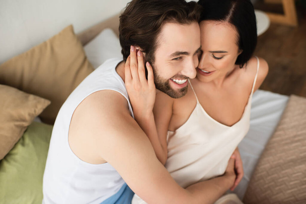 Blick auf die glückliche Freundin im Spaghettikleid, die ihren fröhlichen Freund im Schlafzimmer umarmt - Foto, Bild