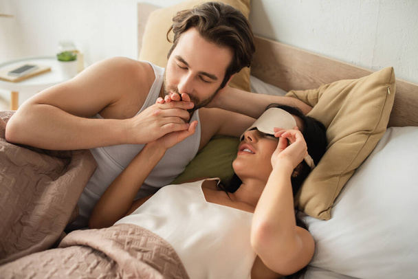 caring man kissing hand of girlfriend in sleeping mask lying in bed - Photo, image