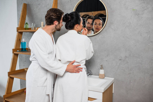 young couple in white bathrobes looking at mirror in bathroom - Фото, изображение
