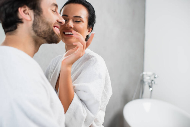 cheerful woman applying face cream on nose of smiling boyfriend in bathrobe - Foto, Bild