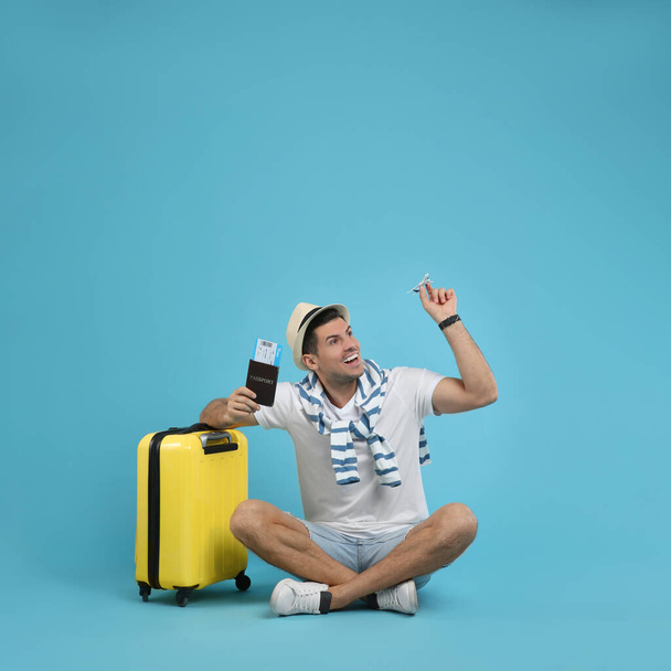 Male tourist holding passport with ticket and toy airplane near suitcase on turquoise background, space for text - Foto, afbeelding