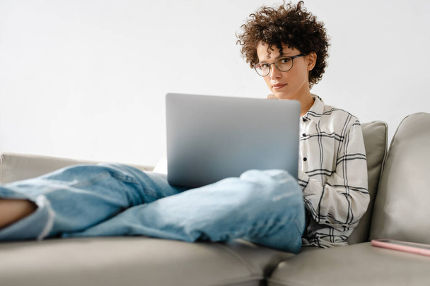 Young curly woman using laptop while sitting on couch at home - Valokuva, kuva