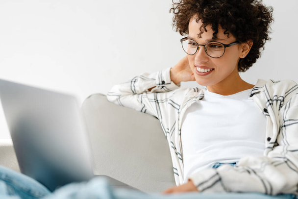 Young curly woman smiling and using laptop while sitting on couch at home - Valokuva, kuva