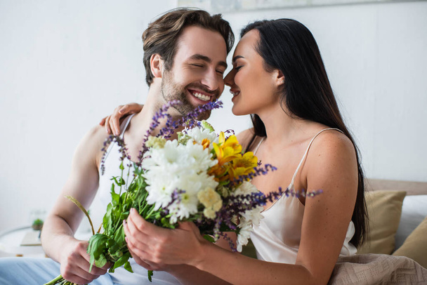 happy man giving bouquet of flowers to smiling girlfriend in bedroom - Photo, Image