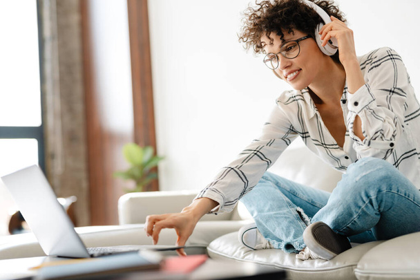 Young curly woman in headphones using laptop while sitting on couch at home - Photo, Image