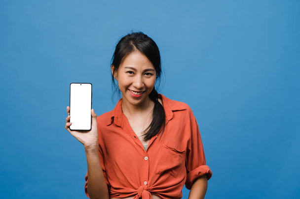 Young Asia lady show empty smartphone screen with positive expression, smiles broadly, dressed in casual clothing feeling happiness on blue background. Mobile phone with white screen in female hand. - Photo, Image