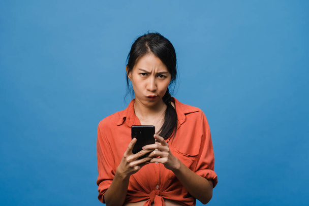 Thinking dreaming young Asia lady using phone with positive expression, dressed in casual clothing feeling happiness and stand isolated on blue background. Happy adorable glad woman rejoices success. - Foto, Imagen