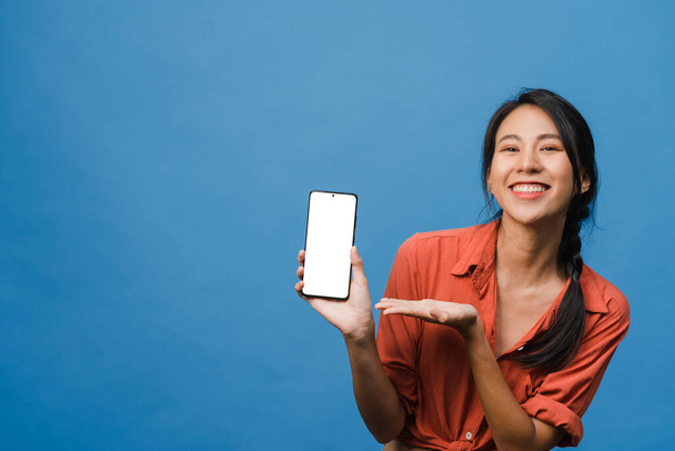 Joven mujer de Asia muestran pantalla de teléfono inteligente vacía con expresión positiva, sonríe ampliamente, vestida con ropa casual sintiendo felicidad en el fondo azul. Teléfono móvil con pantalla blanca en mano femenina. - Foto, imagen