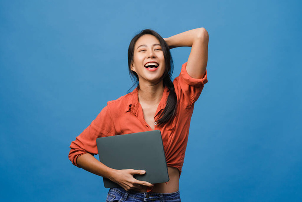 Surprised young Asia lady hold laptop with positive expression, smile broadly, dressed in casual clothing and looking at camera on blue background. Happy adorable glad woman rejoices success. - 写真・画像