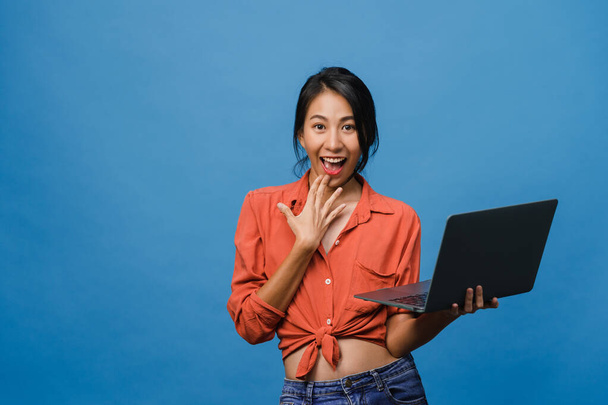 Meglepett fiatal ázsiai hölgy használ laptop pozitív kifejezés, mosoly széles körben, öltözött alkalmi ruházat és nézi a kamera kék háttérrel. Boldog imádnivaló boldog nő örül a sikernek. - Fotó, kép