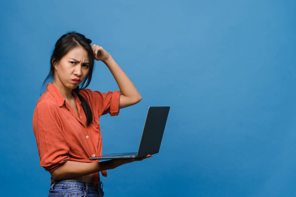 Young Asia lady using laptop with negative expression, excited screaming, cry emotional angry in casual cloth and stand isolated on blue background with blank copy space. Facial expression concept. - 写真・画像