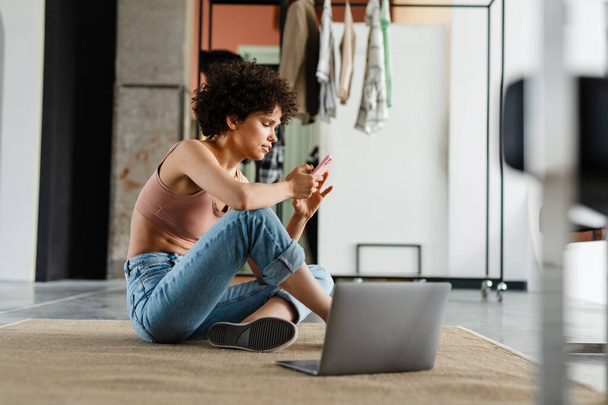 White young woman using mobile phone while sitting on a floor with laptop computer in the modern living room - Photo, image