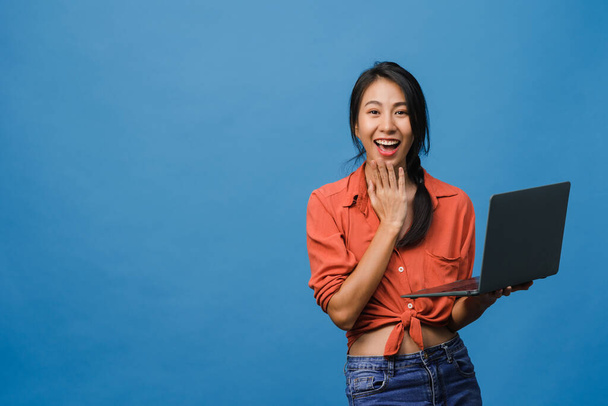 Surprised young Asia lady using laptop with positive expression, smile broadly, dressed in casual clothing and looking at camera on blue background. Happy adorable glad woman rejoices success. - 写真・画像