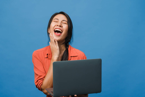 Młoda Azjatka używająca laptopa z pozytywnym wyrazem twarzy, uśmiecha się szeroko, ubrana w luźną odzież, odczuwa szczęście i stoi odizolowana na niebieskim tle. Szczęśliwa, urocza kobieta cieszy się sukcesem.. - Zdjęcie, obraz