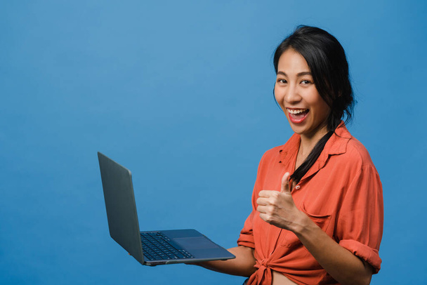 Genç Asyalı bayan laptopunu olumlu ifade ile kullanıyor, geniş bir şekilde gülümsüyor, günlük giysiler içinde mutluluğu hissediyor ve mavi arka planda tek başına duruyor. Mutlu, mutlu, mutlu kadın başarıya sevinir.. - Fotoğraf, Görsel