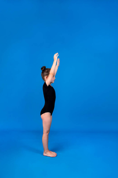 a little girl in a black swimsuit performs a warm-up on a blue background with a place for text - Photo, Image