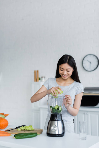 happy asian woman adding sliced cucumbers into shaker near fresh vegetables and chopping board - Photo, Image