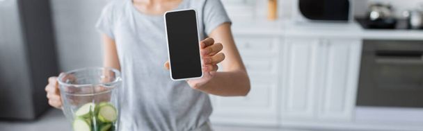 bijgesneden weergave van wazig vrouw met smartphone met leeg scherm in de buurt van shaker met courgette, banner - Foto, afbeelding