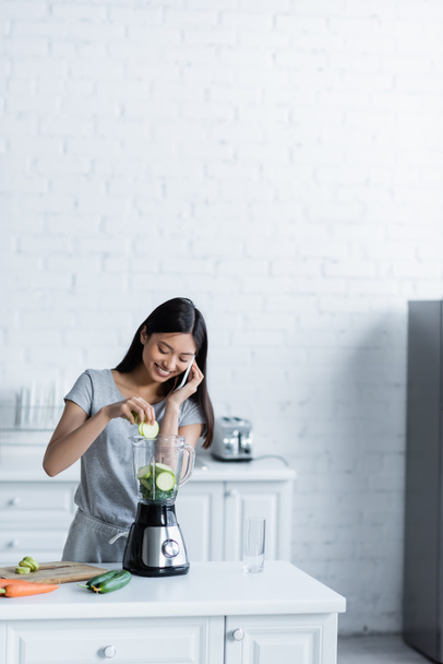 happy asian woman talking on mobile phone while preparing vegetable smoothie in blender - Fotografie, Obrázek