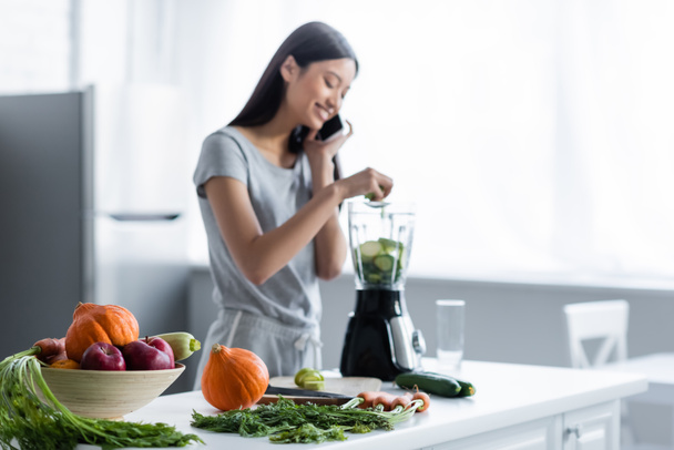 blurred asian woman talking on cellphone near fresh vegetables and electric shaker in kitchen - Photo, Image