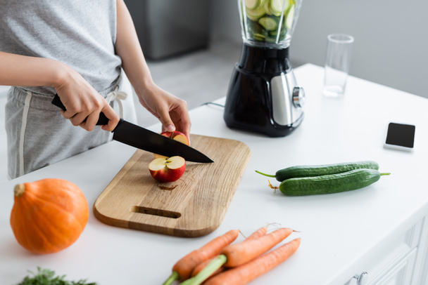 vista parcial de la mujer cortando manzana cerca de calabaza, pepinos y zanahorias en la mesa de la cocina - Foto, imagen