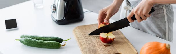 cropped view of woman cutting apple near fresh cucumbers and electric blender, banner - 写真・画像
