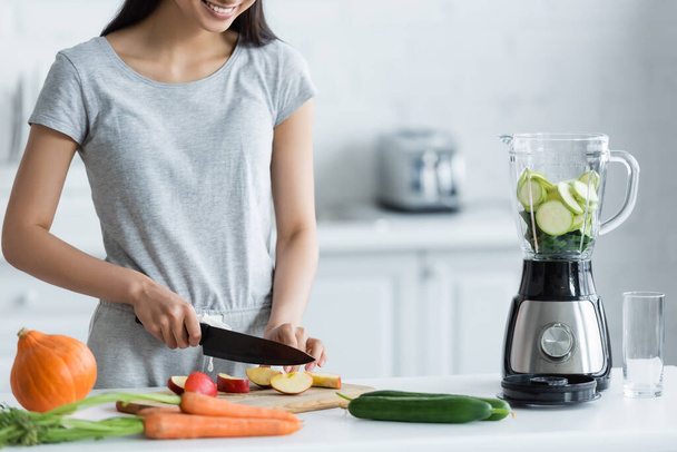 cropped view of happy asian woman cutting apple near electric blender and fresh vegetables on kitchen table - Photo, Image