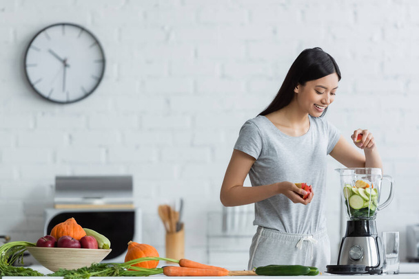 happy asian woman adding apple into shaker with sliced zucchini near fresh vegetables in kitchen - Zdjęcie, obraz