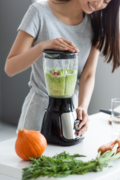partial view of smiling asian woman preparing smoothie near raw pumpkin and carrots - Photo, Image