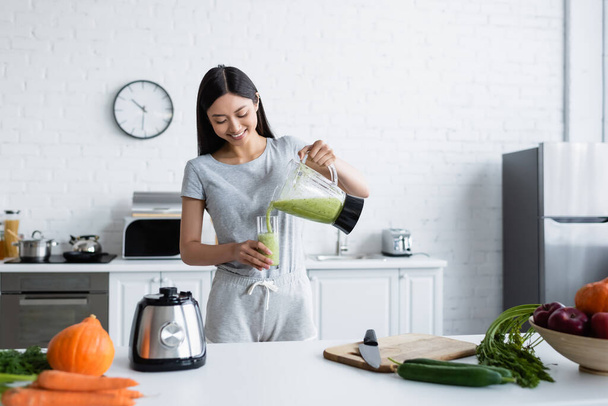 smiling asian woman pouring homemade smoothie near fresh vegetables in kitchen - Φωτογραφία, εικόνα