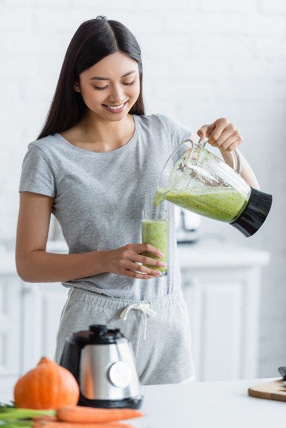cheerful asian woman pouring homemade smoothie into glass near blurred vegetables in kitchen - Fotó, kép
