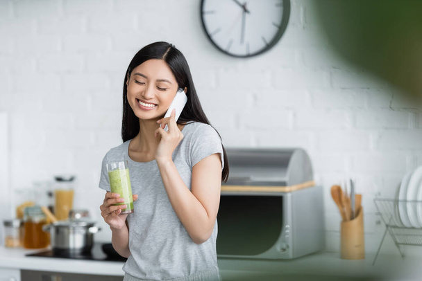 sonriente asiático mujer con vidrio de fresco smoothie hablando en celular en cocina - Foto, imagen