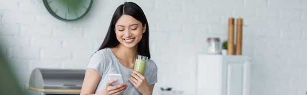 smiling asian woman with glass of delicious smoothie using smartphone in kitchen, banner - Fotó, kép