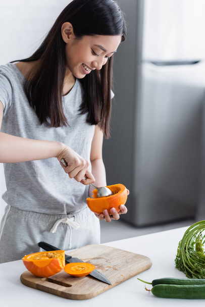 smiling asian woman cleaning out ripe pumpkin near fresh cucumbers on table - Zdjęcie, obraz