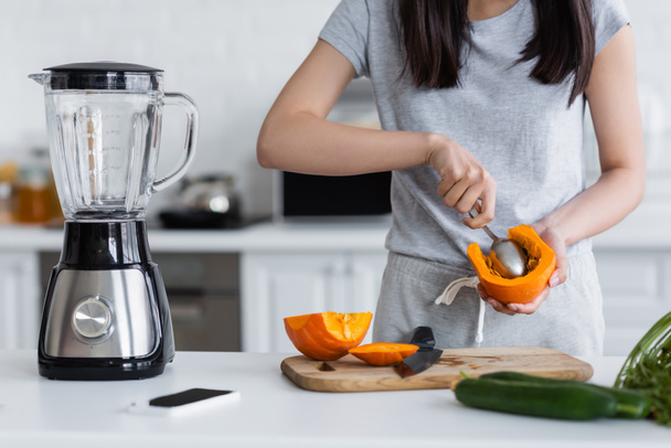partial view of woman cutting out pumpkin with spoon near electric shaker, smartphone and blurred cucumbers - Photo, Image