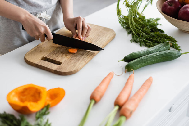 partial view of woman cutting carrot near fresh cucumbers, carrots and pumpkin on table - Foto, Imagem