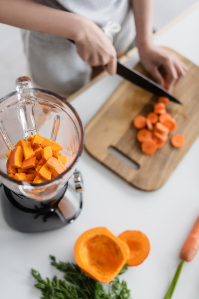 partial view of blurred woman cutting carrot near blender with pumpkin - Foto, imagen
