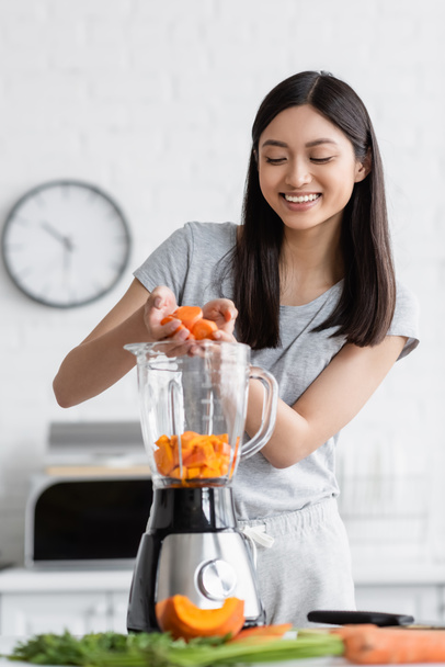 cheerful asian woman adding sliced carrot into electric blender with cut pumpkin - Φωτογραφία, εικόνα