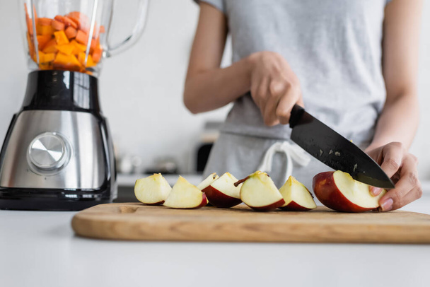 cropped view of blurred woman cutting apple on chopping board near electric shaker - Фото, изображение