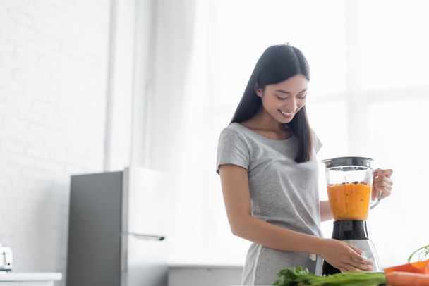 cheerful asian woman preparing vegetable smoothie for breakfast in kitchen - Photo, Image