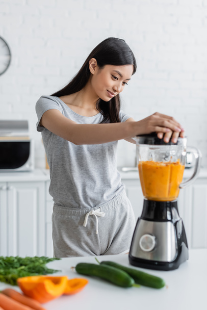 young asian woman preparing fresh smoothie in blender near blurred vegetables on table - Photo, Image