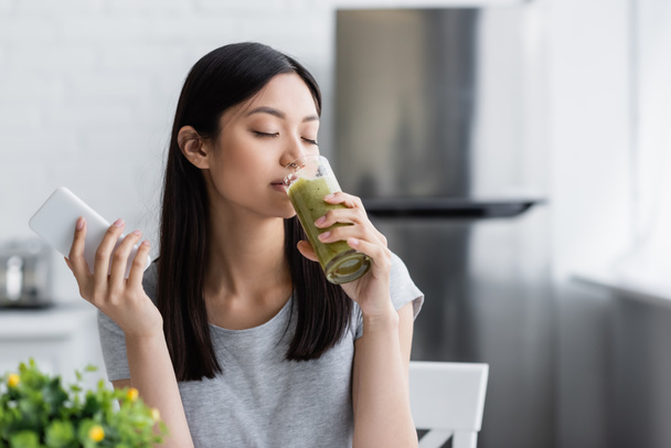 pleased asian woman with mobile phone drinking delicious smoothie in kitchen - Photo, Image