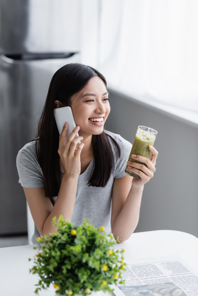 χαρούμενη Ασιάτισσα γυναίκα με ένα ποτήρι φρέσκο smoothie να μιλάει στο κινητό τηλέφωνο κοντά στο θολή φυτό - Φωτογραφία, εικόνα