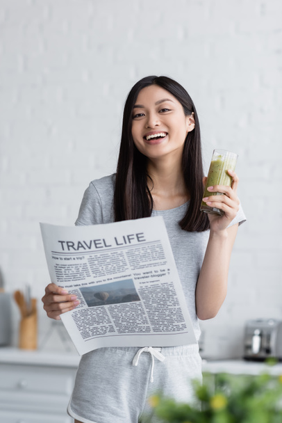 szczęśliwy Azji kobieta z podróż życie gazeta i szklanka świeże smoothie patrząc na aparat fotograficzny w kuchni - Zdjęcie, obraz
