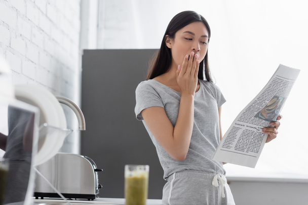 somnoliento asiático chica bostezando mientras leer mañana periódico en cocina  - Foto, Imagen