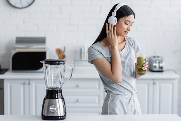 pleased asian woman with glass of smoothie listening music in headphones near electric shaker - Фото, зображення