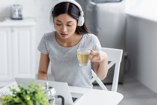  νεαρή ασιάτισσα με ακουστικά κοιτάζοντας το φορητό υπολογιστή, ενώ κρατώντας ένα φλιτζάνι τσάι - Φωτογραφία, εικόνα