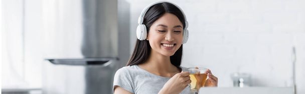felice donna asiatica che tiene in mano una tazza di tè e ascolta musica in cuffia, banner - Foto, immagini