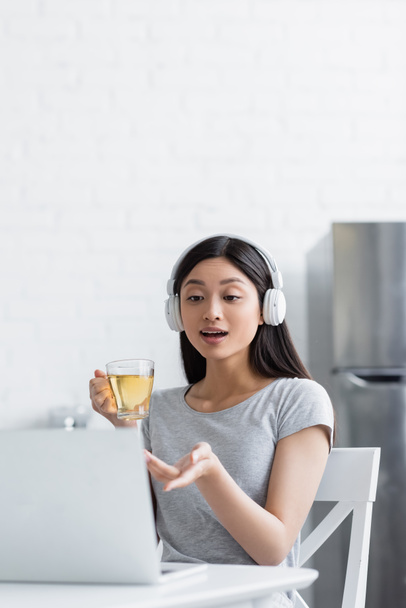 étonné asiatique femme dans casque pointant vers ordinateur portable tout en tenant tasse de thé pf - Photo, image