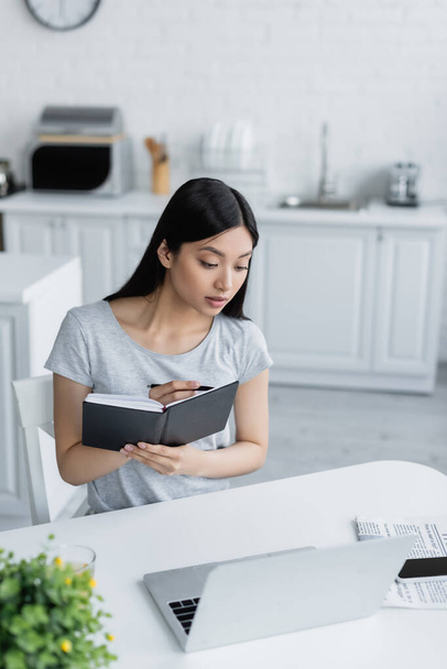 young asian woman writing in notebook near laptop in kitchen - Photo, image